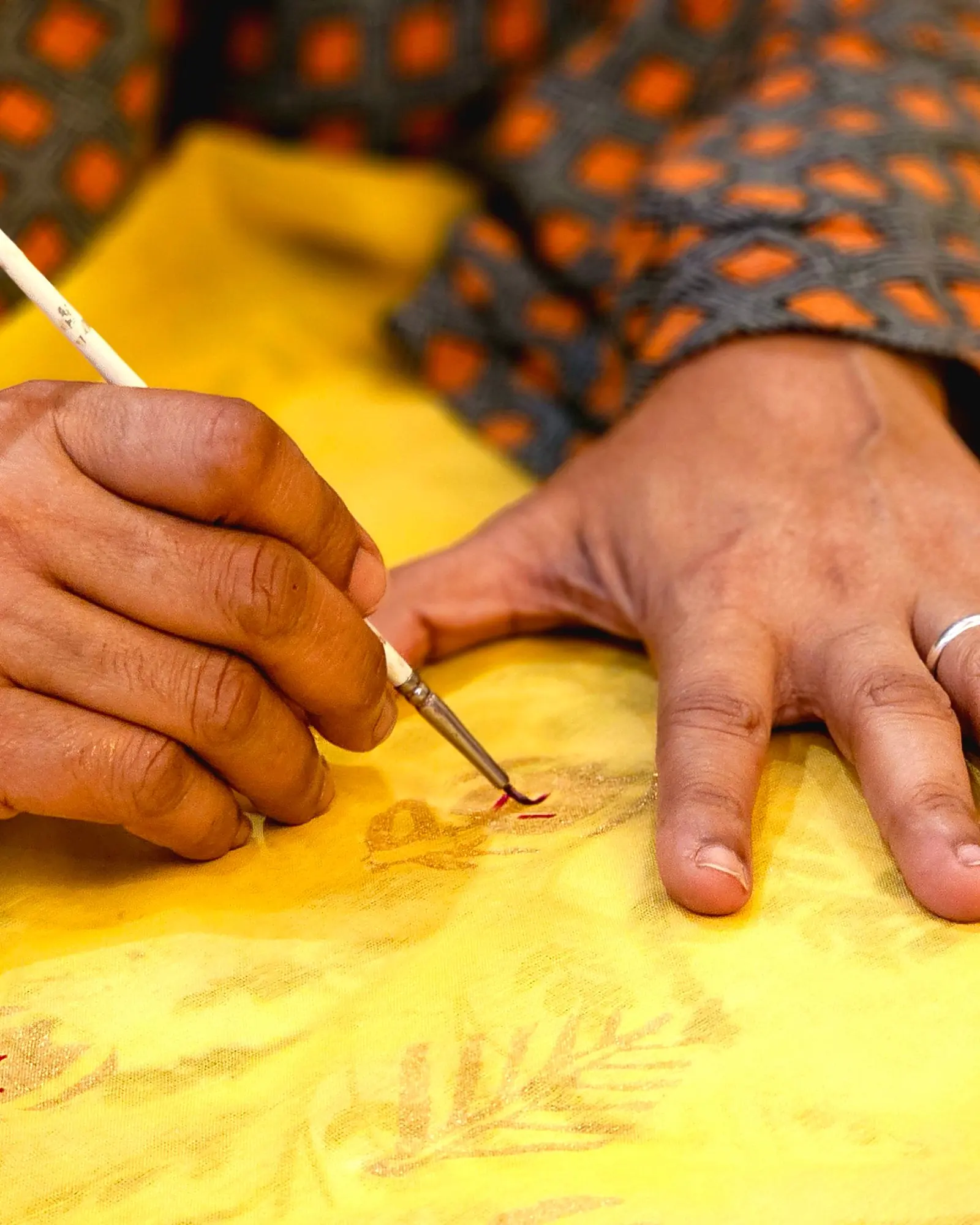 A fine brush held mid-stroke, its tip drawing a dark line into yellow cloth, the other hand spread flat to steady the fabric