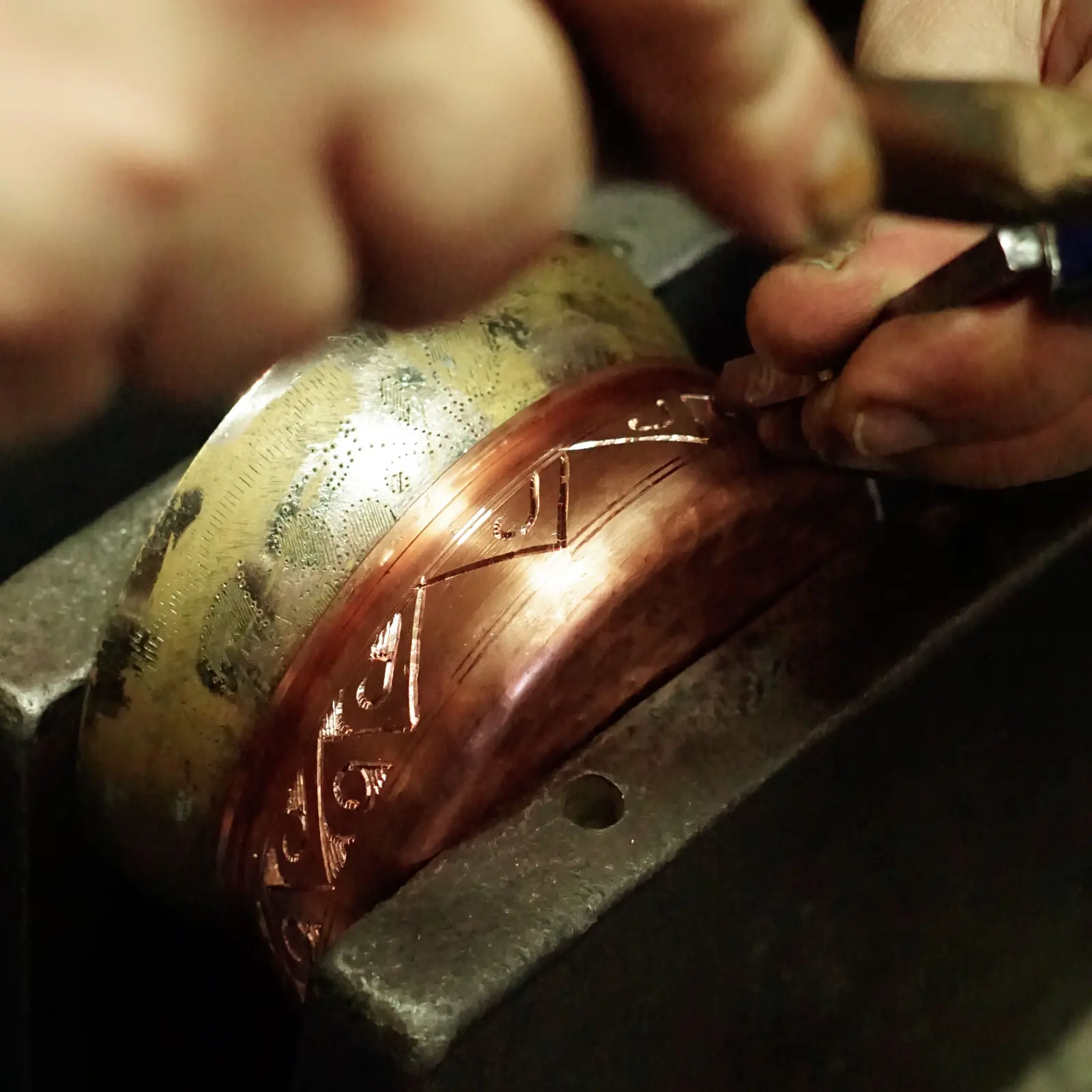 A graver cutting a chevron pattern into a copper bangle held against an anvil, a stippled brass one beside it