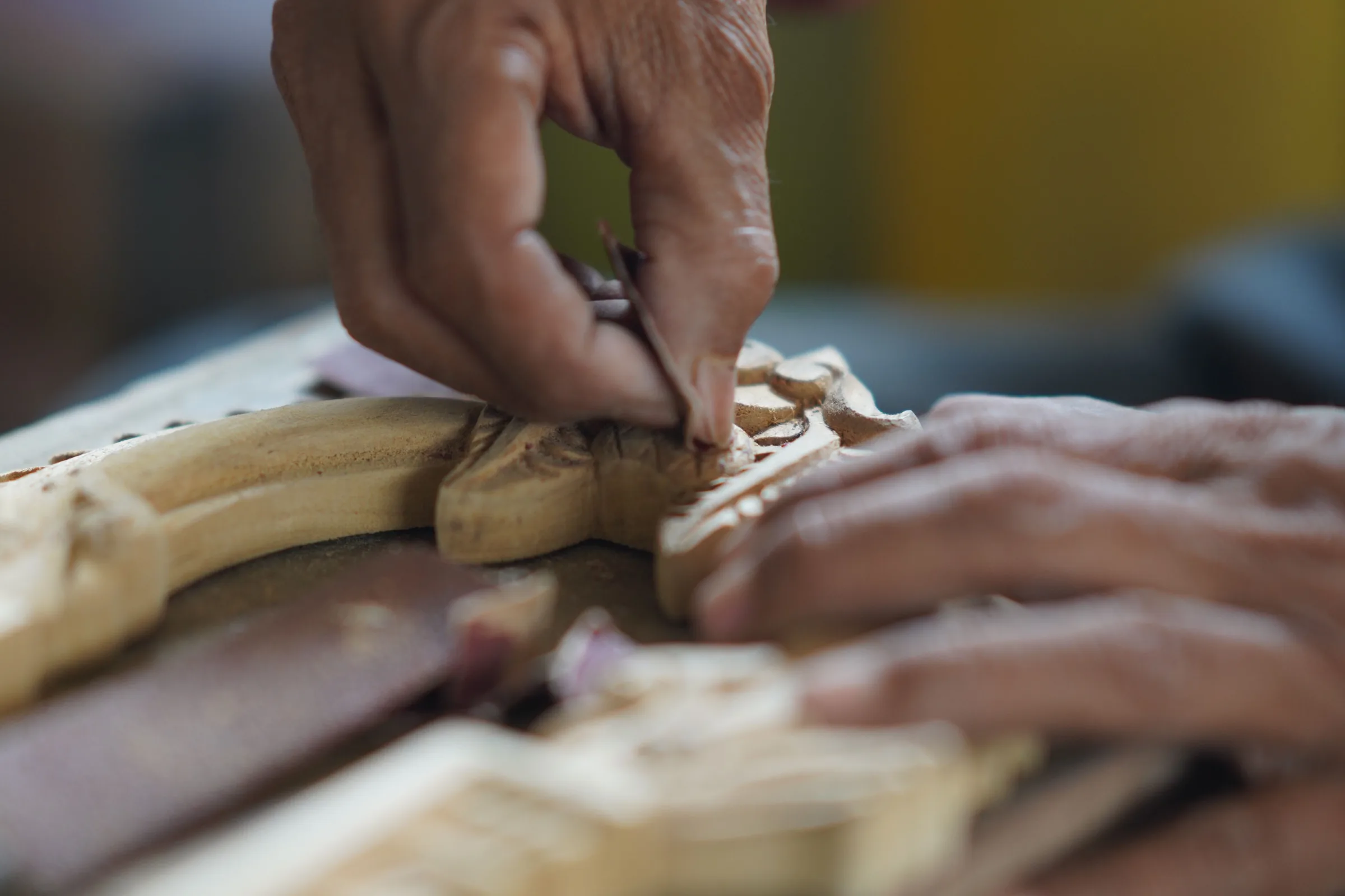 A carver's hands guiding a chisel through a pale wooden panel, shavings gathered around the cut