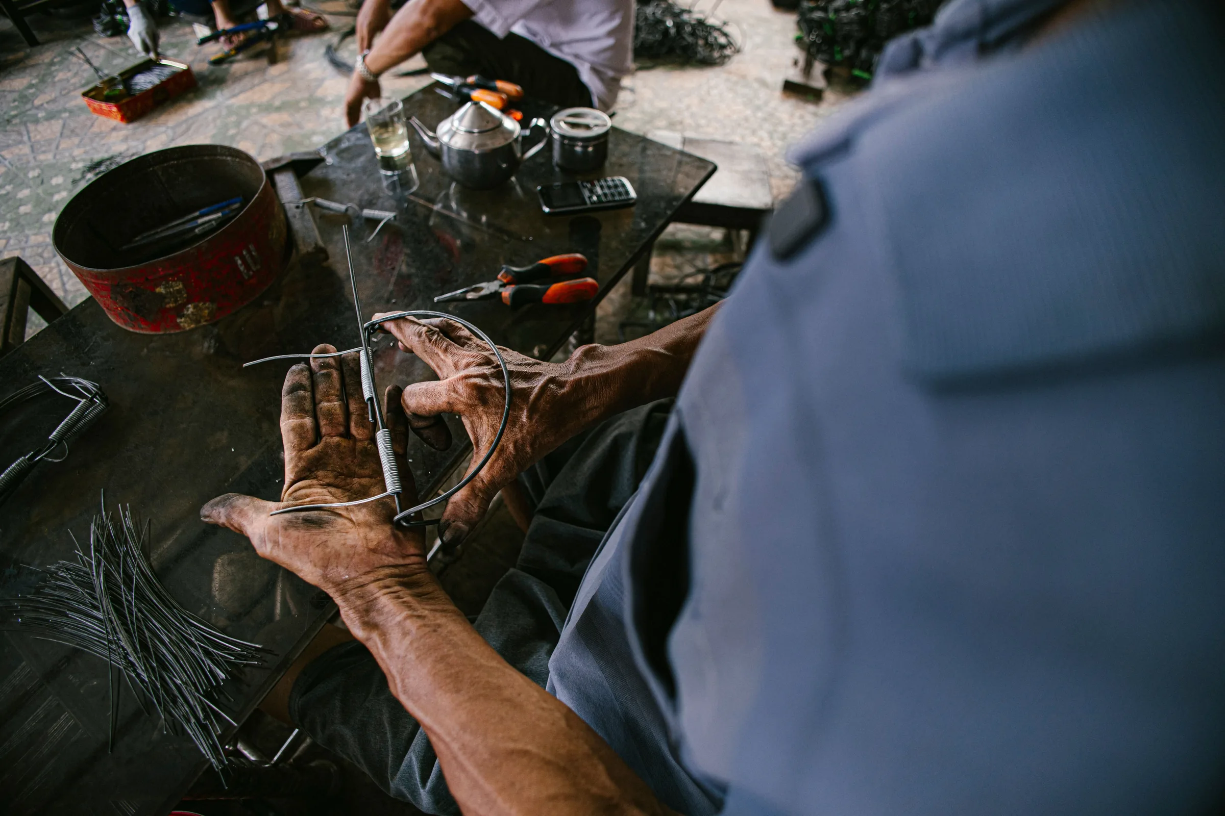 Weathered hands working metal with pliers at a low bench crowded with tools