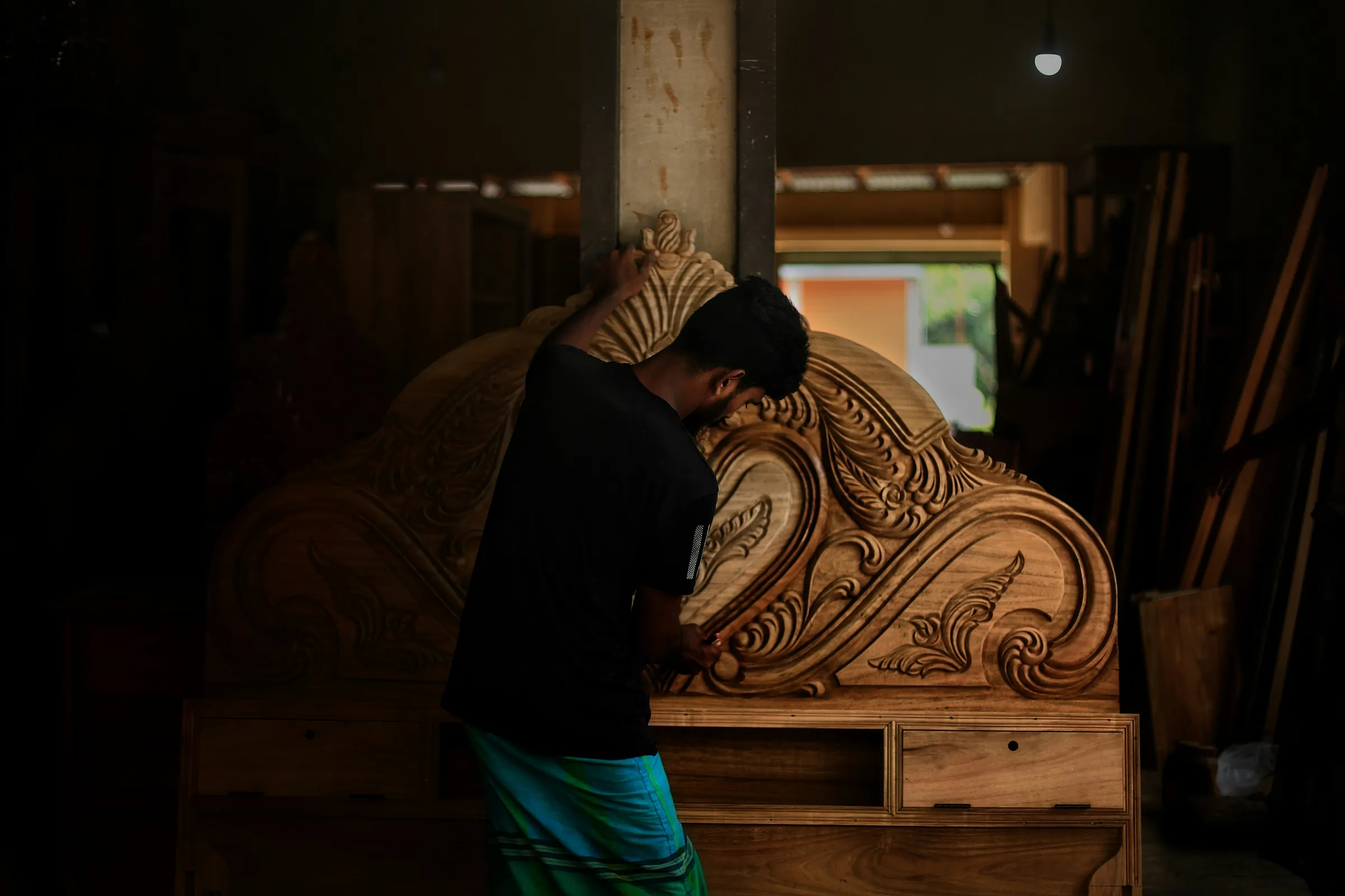 A carver bent close over a wooden headboard in a dim workshop, cutting a scrolling leaf pattern into the pale wood