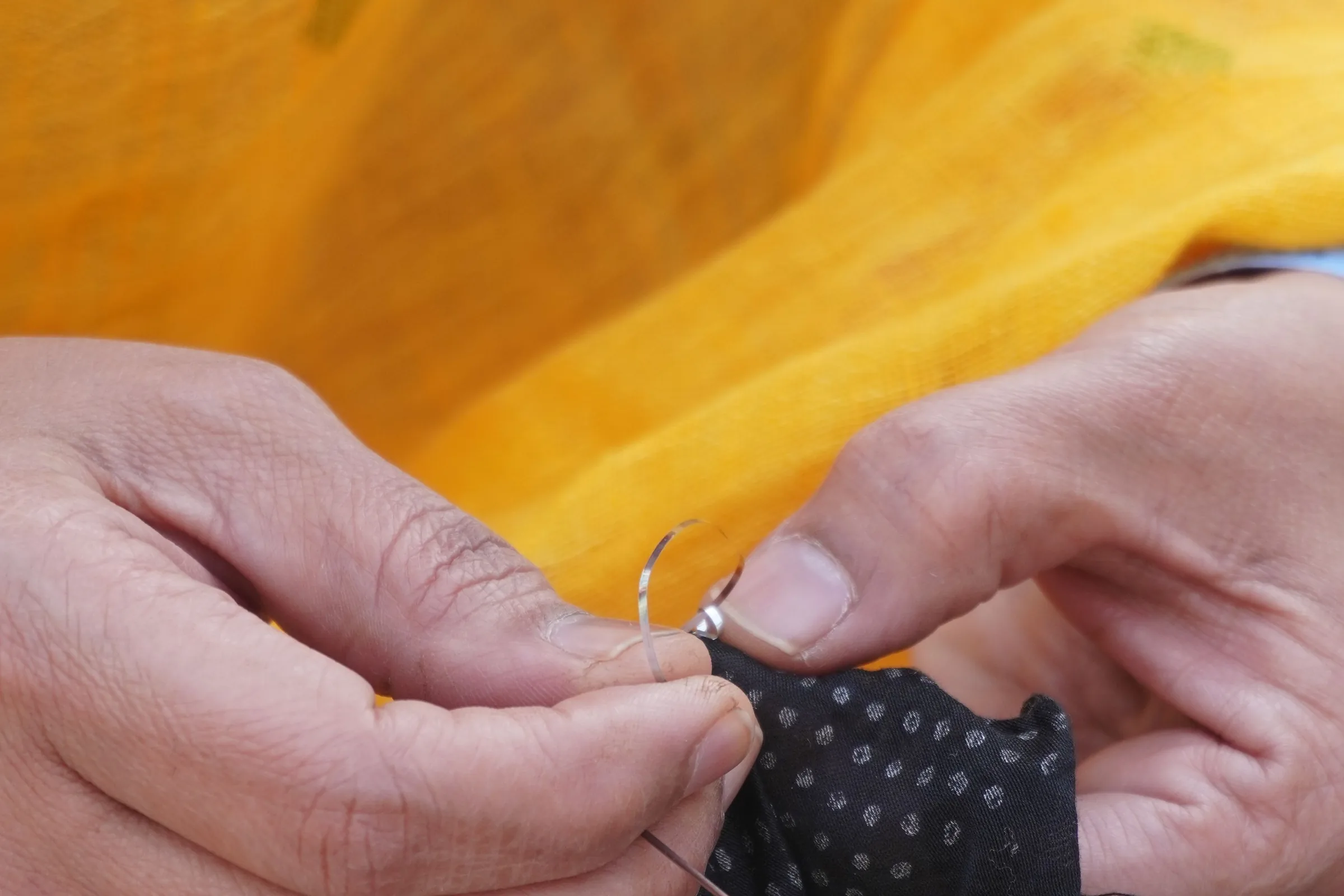 Two hands drawing a needle through dark spotted cloth, against a fold of saffron fabric